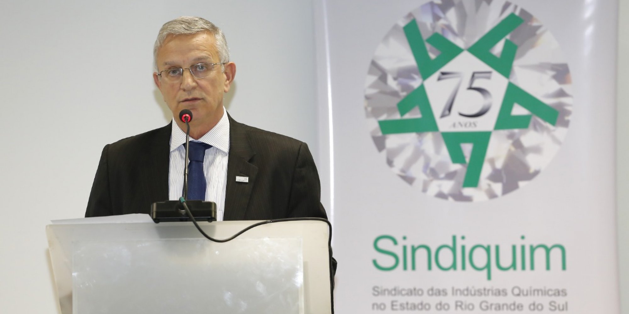 Comemoração dos 75 anos do Sindiquim Homem branco, de óculos, em um púlpito com microfone, discursando, banner do Sindiquim Branco escrito em verde